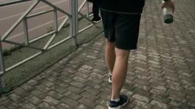 Siyah tişörtlü, şortlu, spor ayakkabılı, spor ayakkabılı, çantalı, kaldırımda yürüyen bir şişe su taşıyan, spor salonuna giderken yolda koşan bir sporcunun orta boy arka çekimi.
