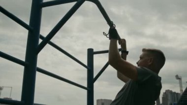 Uzuv farkı olan, protez el kancası takmış, stadyumda hava bulutlu iken egzersiz yaparken barfiks çeken kararlı genç sporcunun orta boy yakın çekimi.