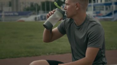 Orta kalibreli, düşünceli, genç, beyaz erkek sporcu. Bir eli engelli, dışarıda stadyumdaki antrenmandan sonra yedek kulübesinde dinleniyor, şişeden su içiyor.