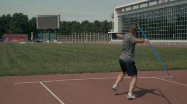 Eksik eli, tişörtü ve şortu olan, pistte koşan, sonra yeşil alana cirit atan ve yaz günü stadyumda tek başına antrenman yapan erkek sporcunun tam arkası.