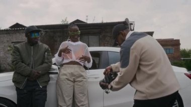 Açık havada müzik videosu kaydetmek için orta uzunlukta bir çekim. Genç erkek kameraman, arabanın yanında kadın ve erkek hip-hop sanatçılarını filme alıyor, bayan, eski püskü bölgede kamera ve kameraman için rap yapıyor.