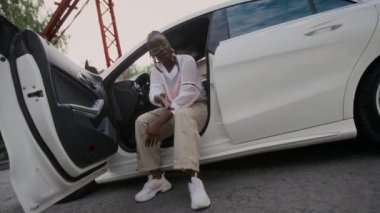 Havalı, genç, Afro-Amerikan hip-hop sanatçısı, şık giyinmiş, gözlüklü, beyaz, modern bir arabada oturan, kapısı açık ve video klip için kameraya vuran bir el kamerasıyla.