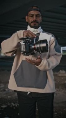Sakallı, bıyıklı, gözlüklü, kepli ve eşofmanlı, şehir merkezinde kamerayla poz veren erkek çift taraflı video fotoğrafçısının dikey portresi.