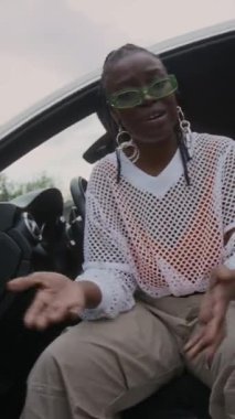 Hip-hop klas kıyafetli, gözlüklü, beyaz, modern bir arabada açık kapılı ve açık havada müzik videosu çekerken kameraya rap yapan arsız Afro-Amerikan kadının dikey düşük açılı görüntüsü.
