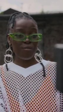 Gözlüklü ve şık giyinmiş genç Afrikalı bir kadının eski püskü bir bölgede müzik videosu çekerken kameraya rap yaptığı el kamerası görüntüsü.