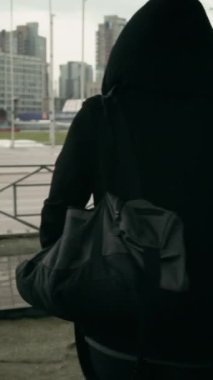 Dikey orta boy siyah kazaklı, spor çanta taşıyan, idman için şehir stadyumuna giden bariyerlerin orada duran ve kapüşonunu çıkartan tanınmamış bir gencin fotoğrafı.