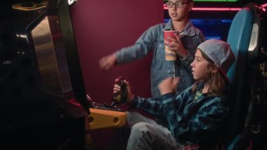 İki çoklu etnik kökenli gencin tatilde atari salonunda takıldığı orta boy bir fotoğraf. Beyaz çocuk kumar makinesinde oyun oynuyor, joystick kullanıyor, Hintli arkadaşı izliyor ve tezahürat yapıyor.