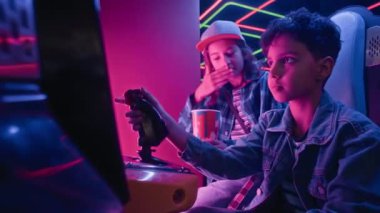 Oyun salonunda oyun oynayan küçük Hintli bir çocuğun orta boy bir video oyunu, beyaz bir arkadaşın izlemesi, yorumlaması, öğüt vermesi, patlamış mısır yemesi, çocuğun kazanması ve beşlik çakması.