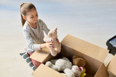 Evin zemininde oturan ve kutuları oyuncak ayılarla boşaltan yüksek açılı küçük bir kız.