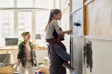 Şirin küçük kız babasının omuzlarında oturuyor ve anne babasına yardım ederken oturma odasının duvarını gri renklerle boyuyor.