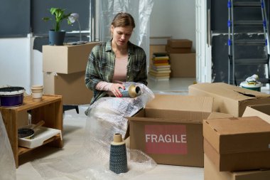 Günlük giysili genç bir kadın, kutuya koymadan önce çömelmiş vazoları baloncuklu ambalaja sarıp, süslüyor.