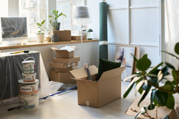 Большая квадратная картонная коробка с бытовой утварью, стоящая на полу гостиной между ведрами с краской и мебелью