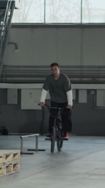 Genç beyaz adamın, BMX bisikletle hangarda sürerken çekilen dikey saplama sahnesi. Temiz bir dönüş yapamamak, arkadaşla pratik yapmak.