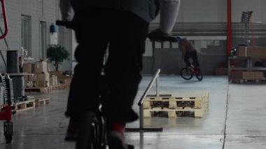 Bisikletli genç BMX sürücünün arka planda çekimleri var. Çelik korkuluklara tırmanıyor ve hangarda dublörlük yaparken zıplıyor. Arka planda dönen numaralar yapıyor.