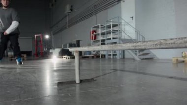 Spor elbiseli, kask takan, çelik raylarda zıplayan ve paten kayan bir gencin antrenman sırasında hangarda paten kayarken çekilmiş yavaş çekimi.