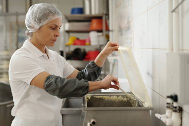 Endüstriyel mutfakta çalışan koruyucu giysili bir kadın, profesyonel bir çevreyi yansıtan paslanmaz çelik bir konteynırda gıda malzemeleri hazırlamaya odaklanmış.