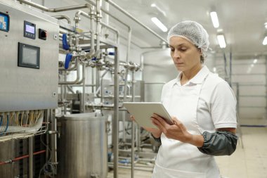 Dijital tablet tutarken steril endüstriyel tesislerdeki makineleri denetleyen mühendis. Parlak, aydınlık bir ortamda saç filesi ve üniforma giyen işçiler.