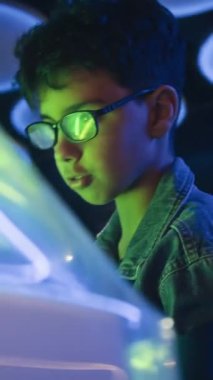 Kot ceketli, sakız çiğneyen, oyun salonunda, neon ışıklarda arkadaşlarıyla masa oyunu oynayan zeki, genç Asyalı çocuğun dikey görüntüsü.