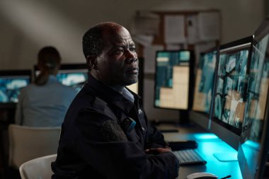 Ciddi bir Afrikalı Amerikalı güvenlik görevlisi gözlem odasında iş yerinde otururken bilgisayar ekranında CCTV videosu üzerinde yoğunlaşıyor.