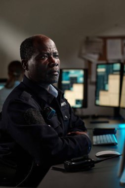 Siyah üniformalı yetişkin erkek güvenlik görevlisi kontrol odasında masanın başında oturuyor ve masaüstü bilgisayar ekranına bakıyor.