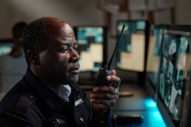 Ciddi bir yetişkin güvenlik görevlisi telsizle durumu bildirirken bilgisayar ekranına bakıyor.