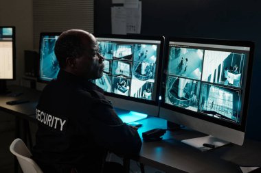 Olgun erkek güvenlik görevlisi gözlem odasındaki birkaç masaüstü bilgisayar ekranından birine bakıyor.