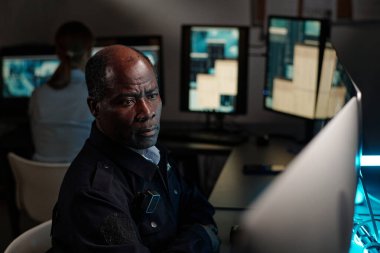 Ciddi bir yetişkin güvenlik görevlisi gözlem odasında iş yerinde oturup masaüstü bilgisayar ekranlarından birine bakıyor.