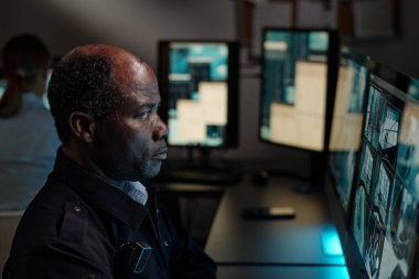 Olgun Afro-Amerikan güvenlik görevlisi izleme odasındaki bilgisayar ekranında güvenlik kamerası görüntülerini izlemeye odaklanıyor.