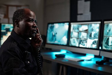 Ciddi bir yetişkin güvenlik görevlisi telefon ahizesi ile iş yerinde bilgisayar ekranlarındaki kapalı devre TV videosunun önünde oturuyor.