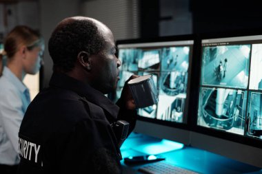 Ciddi bir yetişkin erkek polis memuru bilgisayar ekranlarının önünde oturup kapalı devre televizyon izlerken bir fincan kahve içiyor.