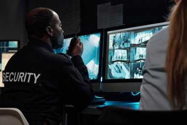 Olgun Afro-Amerikalı adam radyo vericisiyle konuşuyor ve CCTV video sistemi ile bilgisayar ekranına bakıyor.