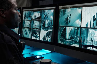 Ciddi bir Afrikalı Amerikalı güvenlik görevlisi güvenlik odasında güvenlik kamerası görüntülerini izlerken bilgisayar ekranından otel hayatını izliyor.