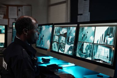 Afrika kökenli yetişkin bir adam iş yerinde monitörlerle otururken bilgisayar ekranına kameralarla bakıyor.