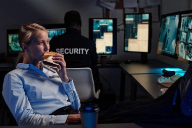 Genç bayan gardiyan iş yerinde bilgisayar monitörü önünde oturup kapalı devre televizyon izlerken sandviç yiyor.