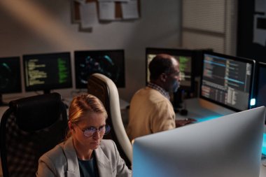 Gözlüklü ve spor ceketli ciddi bir kadın programcı iş arkadaşına karşı veri çözerken bilgisayar ekranına bakıyor.