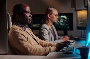 Ciddi bir Afrikalı Amerikalı bilişim mühendisi bilgisayar ekranına bakıyor ve kadın meslektaşının karşısında otururken verileri deşifre ediyor.