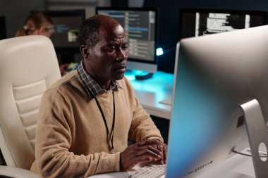 Deneyimli erkek IT destek yöneticisi masaüstü bilgisayar monitörü önünde otururken verileri çözüyor ve ekrana bakıyor