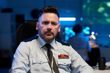 Açık mavi üniformalı, ciddi, olgun bir erkek subay askeri komuta merkezinde çalışırken kameraya bakıyor.