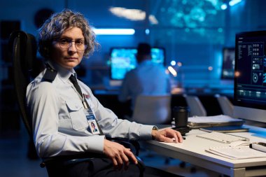 Gözlüklü ve üniformalı deneyimli bayan memur bilgisayar monitörüyle masada otururken kameraya bakıyor.