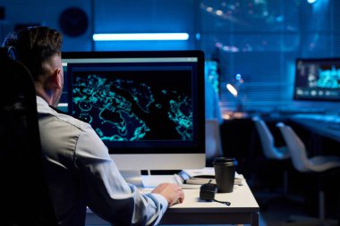 Omzunda, kafasının ve sırtının bir kısmı üniformalı bir erkek memurun masaüstü bilgisayarının önünde elinde verilerle oturuyor.