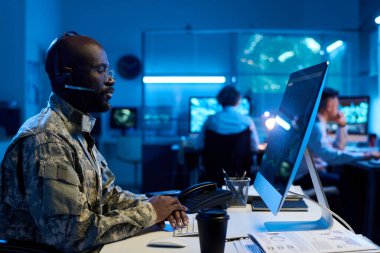 Masaüstü bilgisayar ekranına bakan kulaklıklı, gözlüklü ve kamuflaj üniformalı genç Afrikalı Amerikalı erkek askeri operatör.
