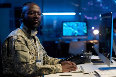 Kamuflaj üniformalı ciddi bir Afrikalı genç adam askeri durumu bildirirken kameraya bakıyor ve daktilo çekiyor.