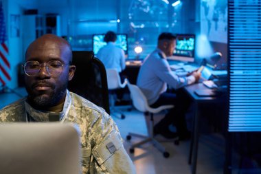 Masaüstü bilgisayarının önünde askeri ofisteki iş arkadaşlarına karşı kamuflaj üniformalı ciddi bir genç adam oturuyor.