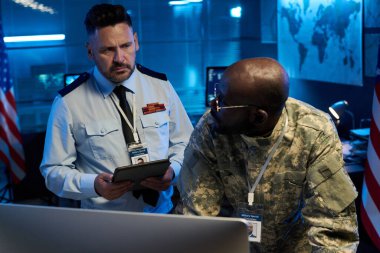 Üniformalı, ciddi bir yetişkin erkek polis memuru bilgisayar monitörüne eğilmiş genç Afrikalı Amerikalı meslektaşına bakıyor.