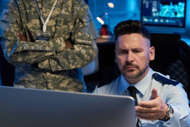 Meslektaşının yanında otururken bilgisayar ekranına yanlış anlaşılmalarla bakan üniformalı ciddi bir olgun adam.