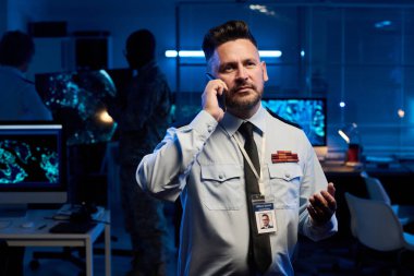 Askeri komuta merkezinde kameranın önünde duran ve cep telefonuyla konuşan kendinden emin olgun bir erkek subay.