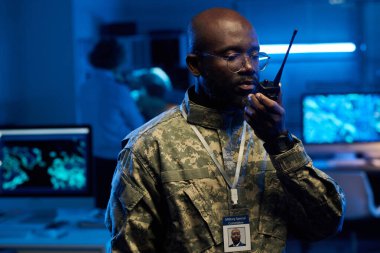 Gözlüklü ve kamuflaj üniformalı genç Afrikalı bir adam kameranın önünde telsizle konuşuyor.