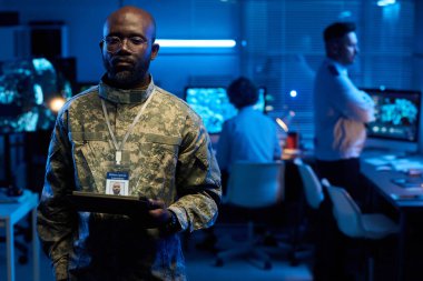 Üniformalı ve gözlüklü ciddi bir genç elinde tabletle askeri ofisteki çevrimiçi verilere bakıyor.