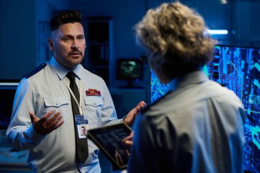 Kendinden emin, olgun bir erkek polis memuru, kadın meslektaşlarının bilgilerini açıklarken, her ikisi de etkileşimli ekranın yanında duruyor.