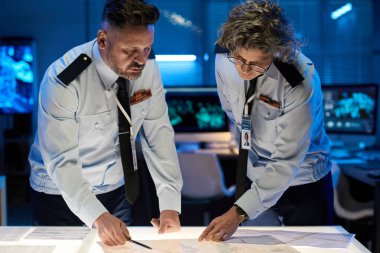 Olgun erkek ve kadın ordu subayları sunum sırasında verilere işaret ederken kağıt belgelerle masanın üzerine eğildiler.